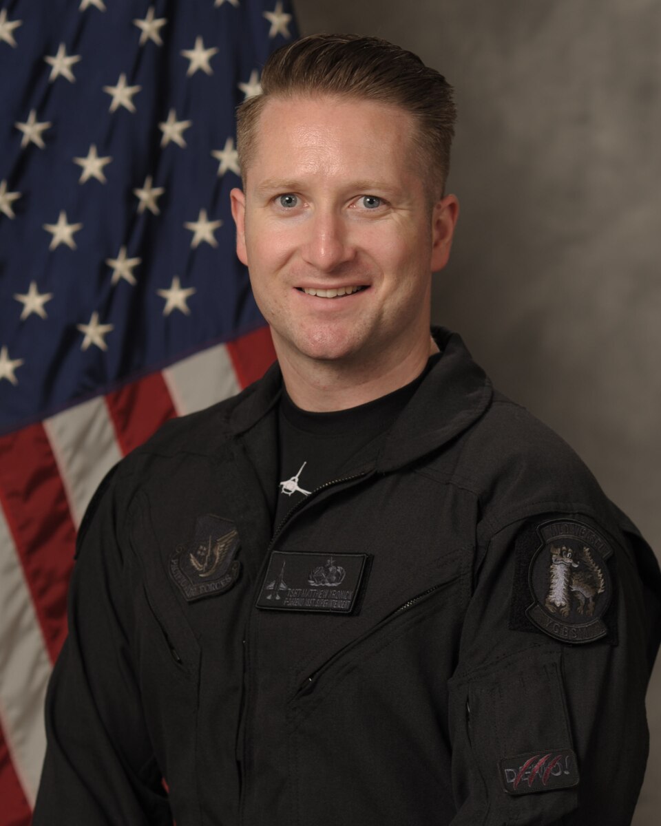 A man in a black flight suit in front of an American Flag.