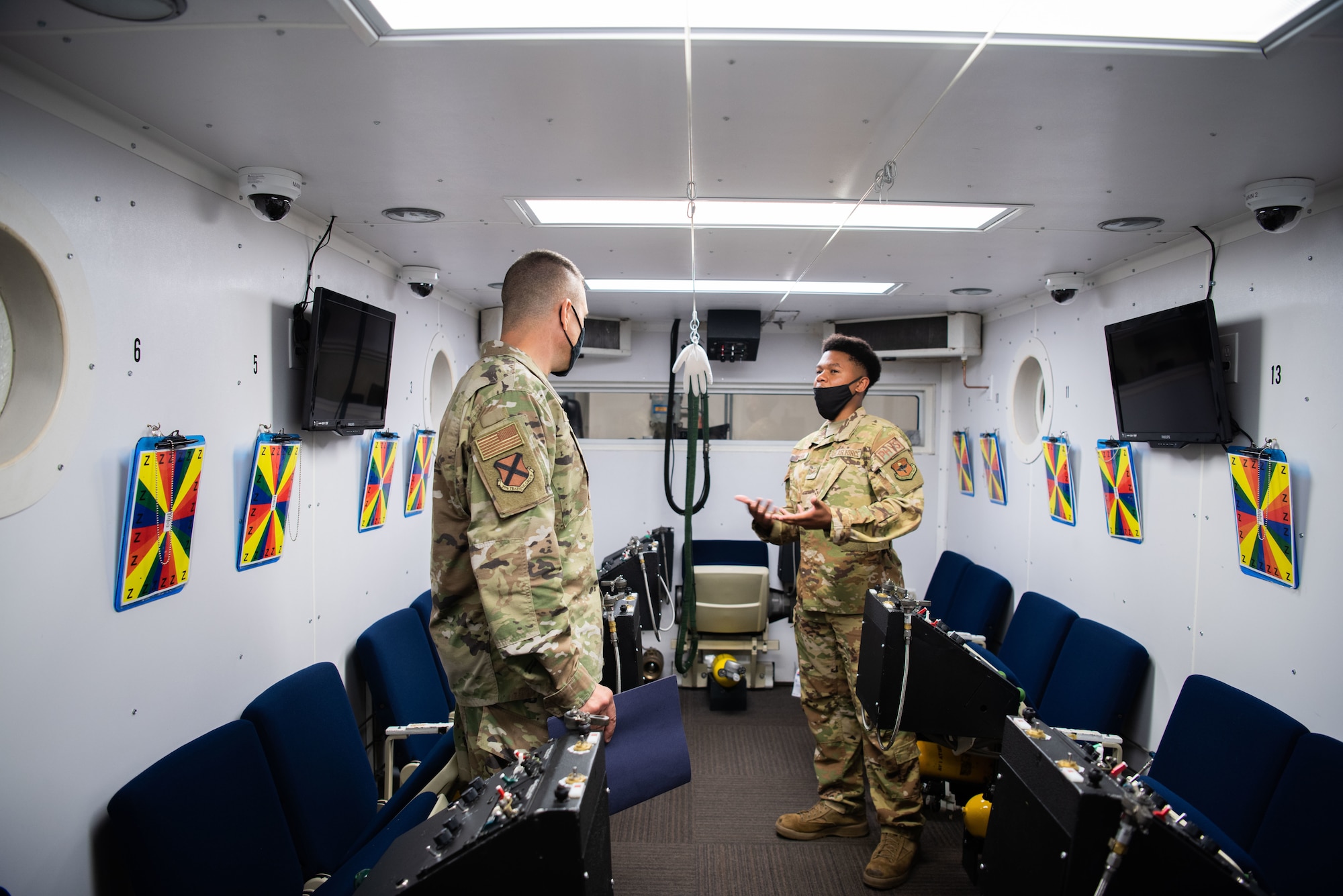 Two men standing and talking inside an altitude chamber. There are rows of chairs and two televisions on the left and right side of the chamber. There are also clipboards hanging on the walls above each chair.