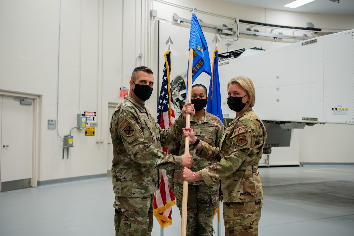Two people holding onto a guidon looking at the camera, and a third person placed in the middle looking straight ahead at the camera. Part of the centrifuge is in the background.