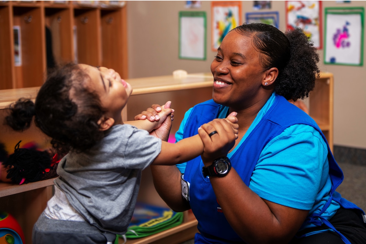 An smiling adult sits on a classroom floor and holds the arms of a child, who leans back and laughs.