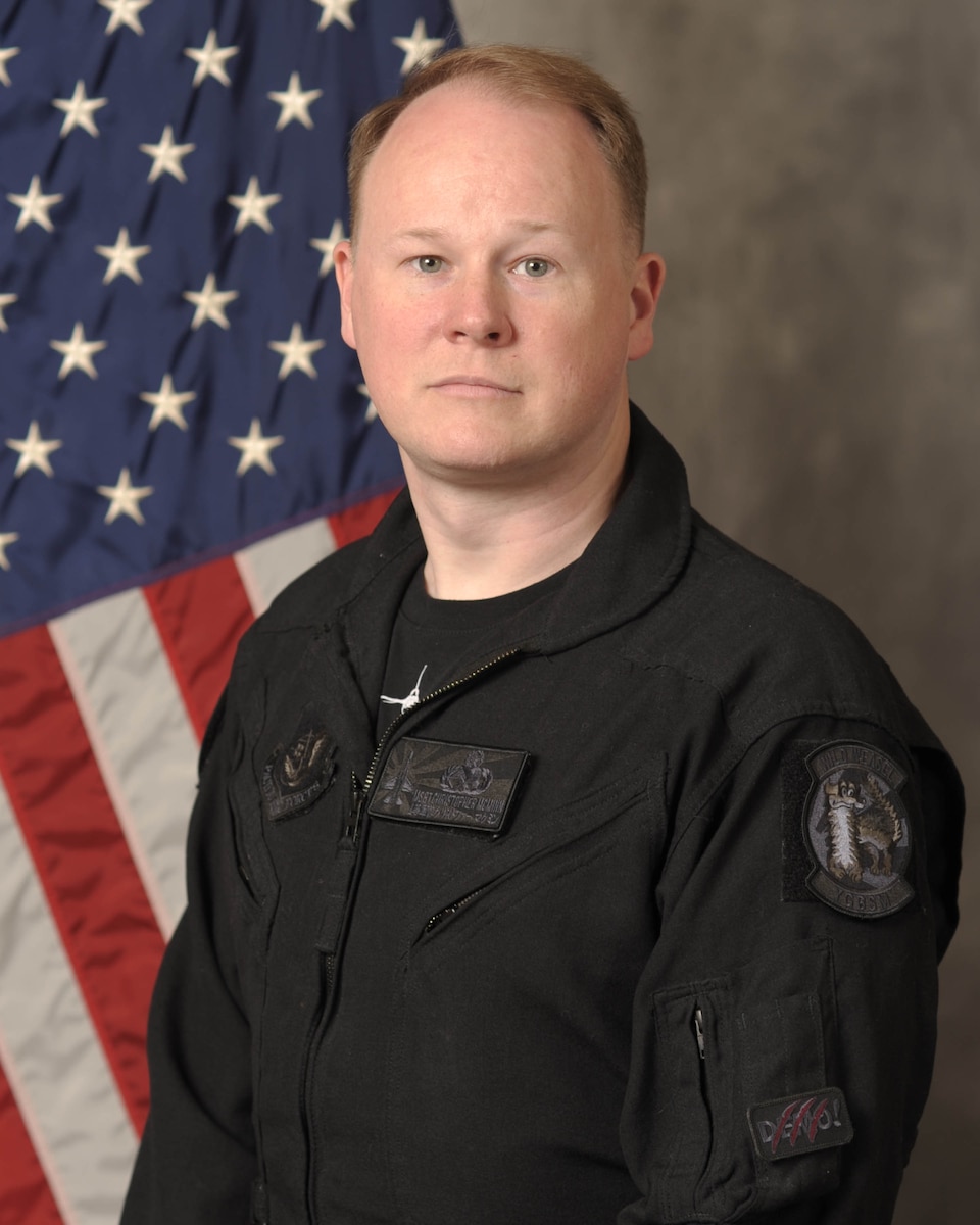 A man in a black flight suit in front of an American flag.