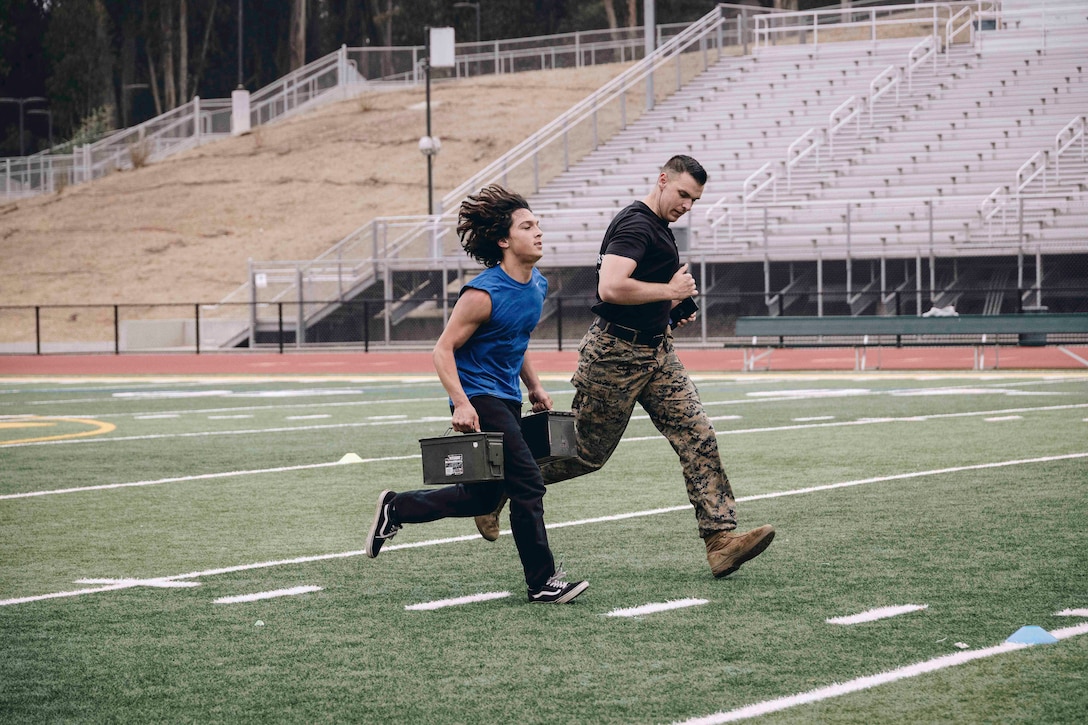 A high school student carrying weighted boxes runs next to a Marine on a field.