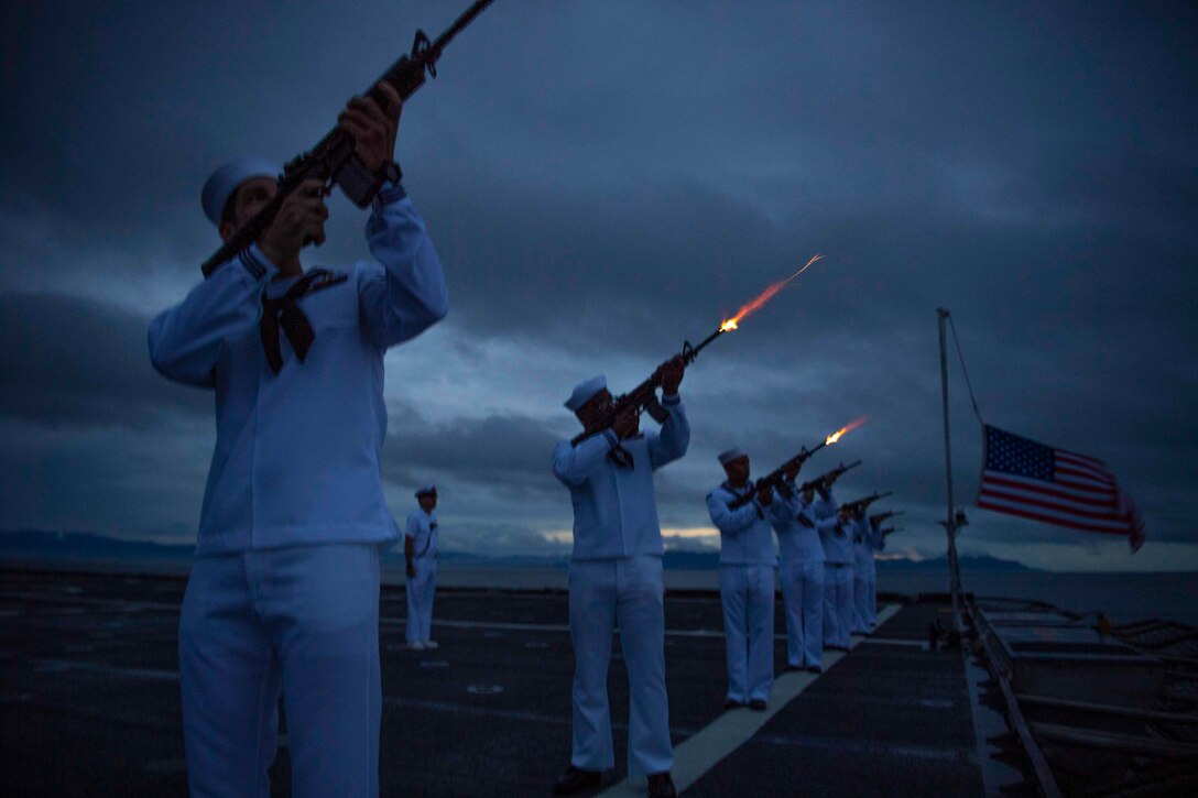 Sailors stand in a line next to an American flag on a ship while firing weapons into a dark sky.