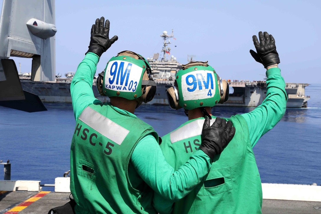 Two sailors wave at a passing aircraft carrier.