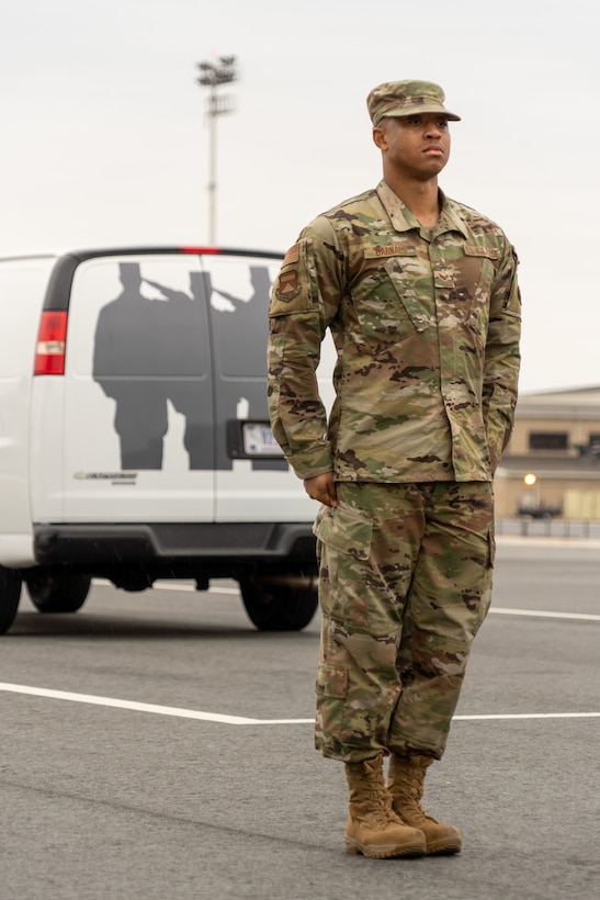 An airman stands at attention in front of a van on a flightline.