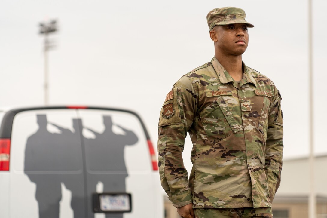 An airman stands at attention in front of a van on a flightline.