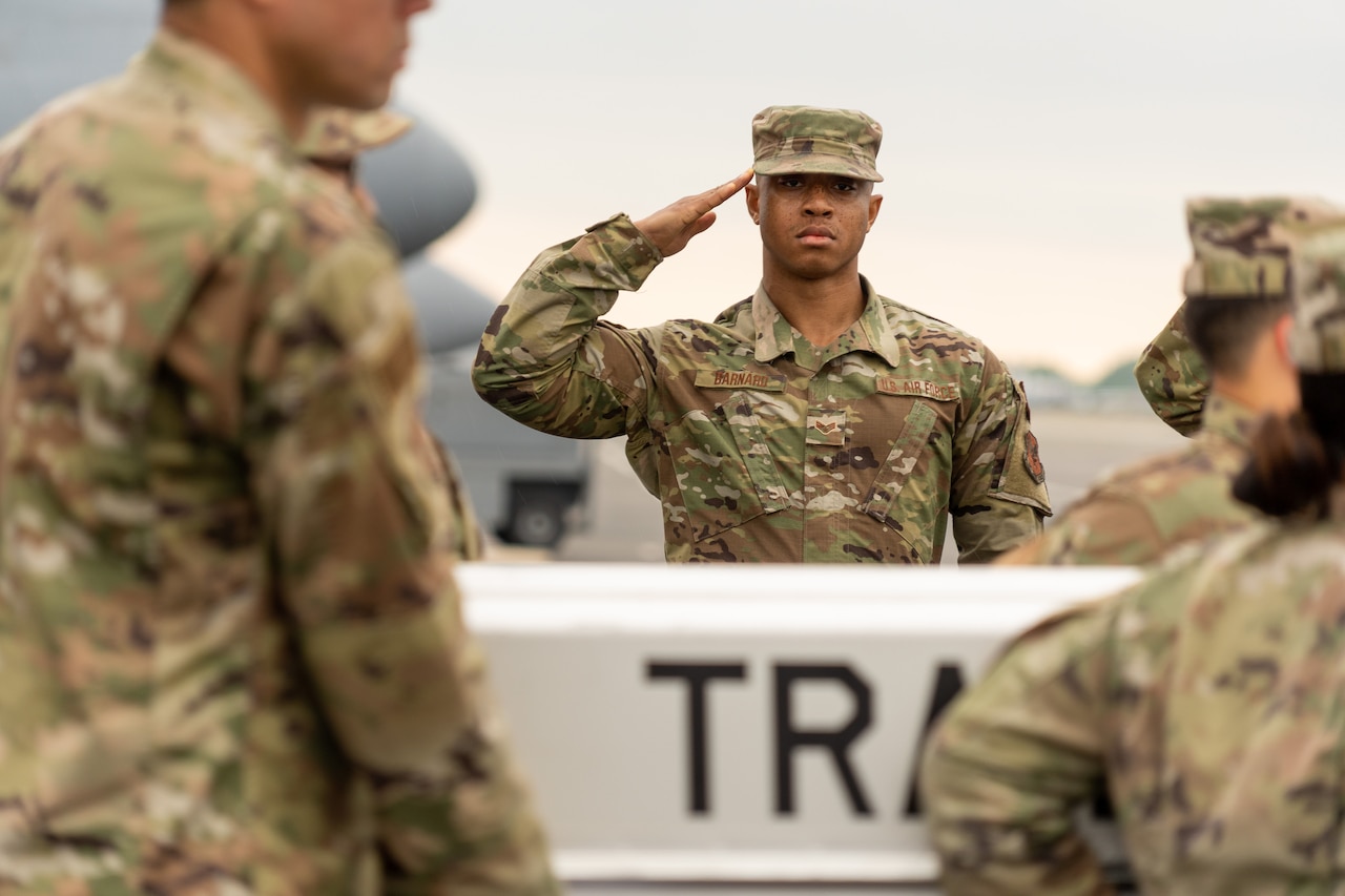 An airman stands outside and salutes as others walk past carrying a case.