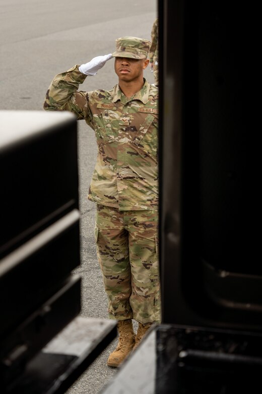 An airman stands outside an open door and salutes wearing a white glove.