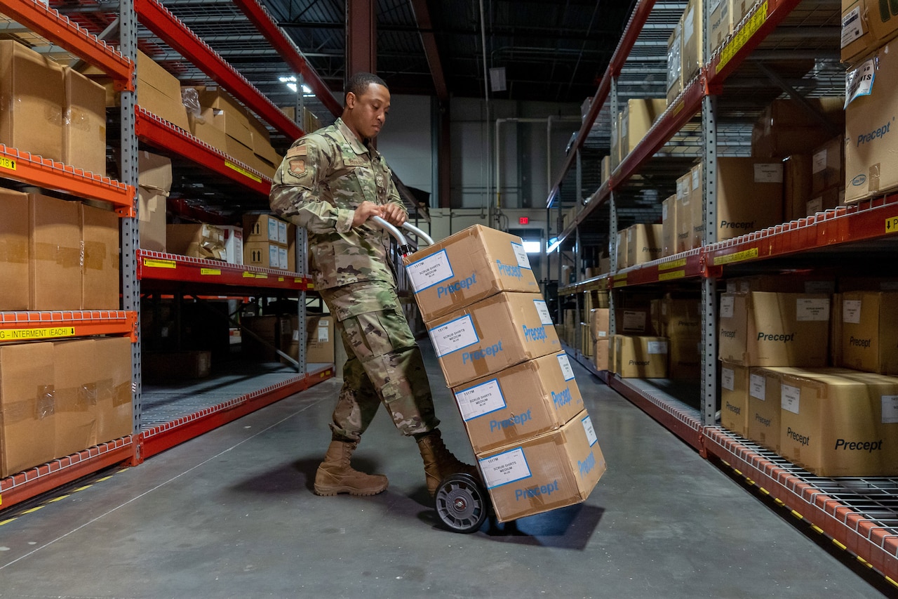 An airman moves cardboard boxes using a dolly in a warehouse-type space.
