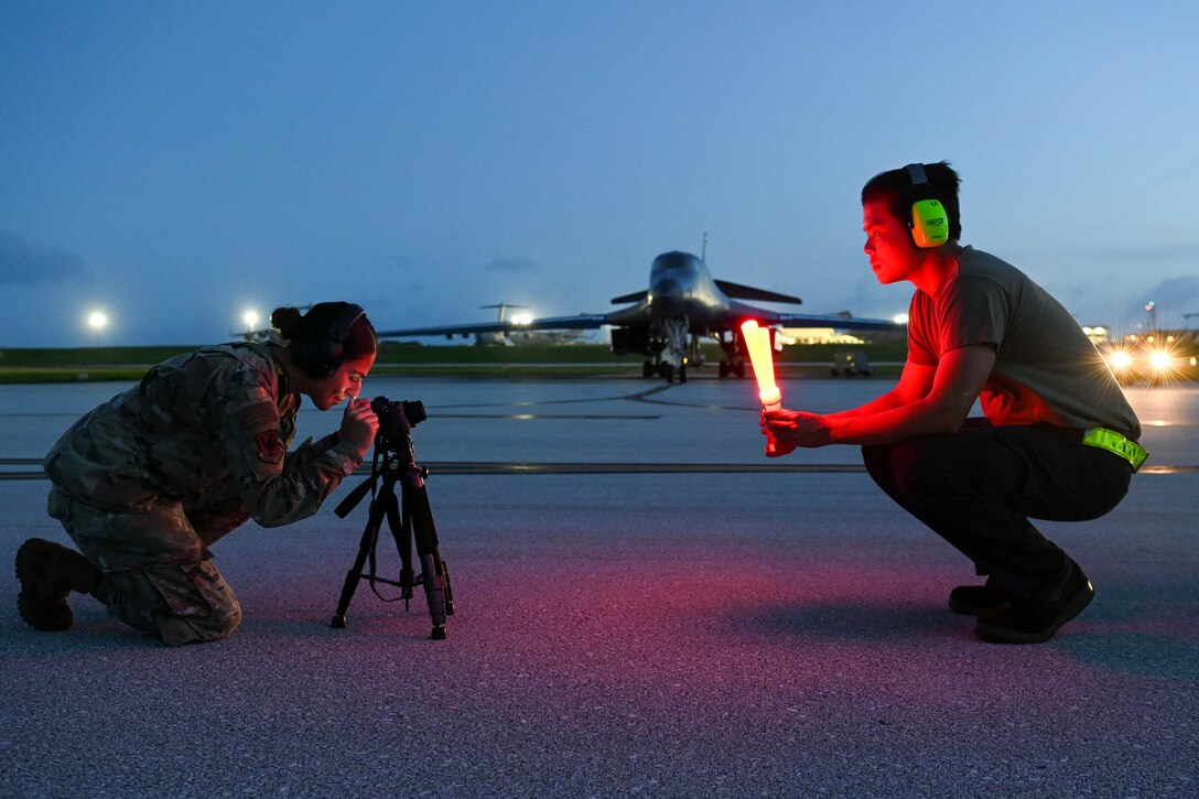 An airman kneels and takes a picture of another airman crouching on a runway at twilight.