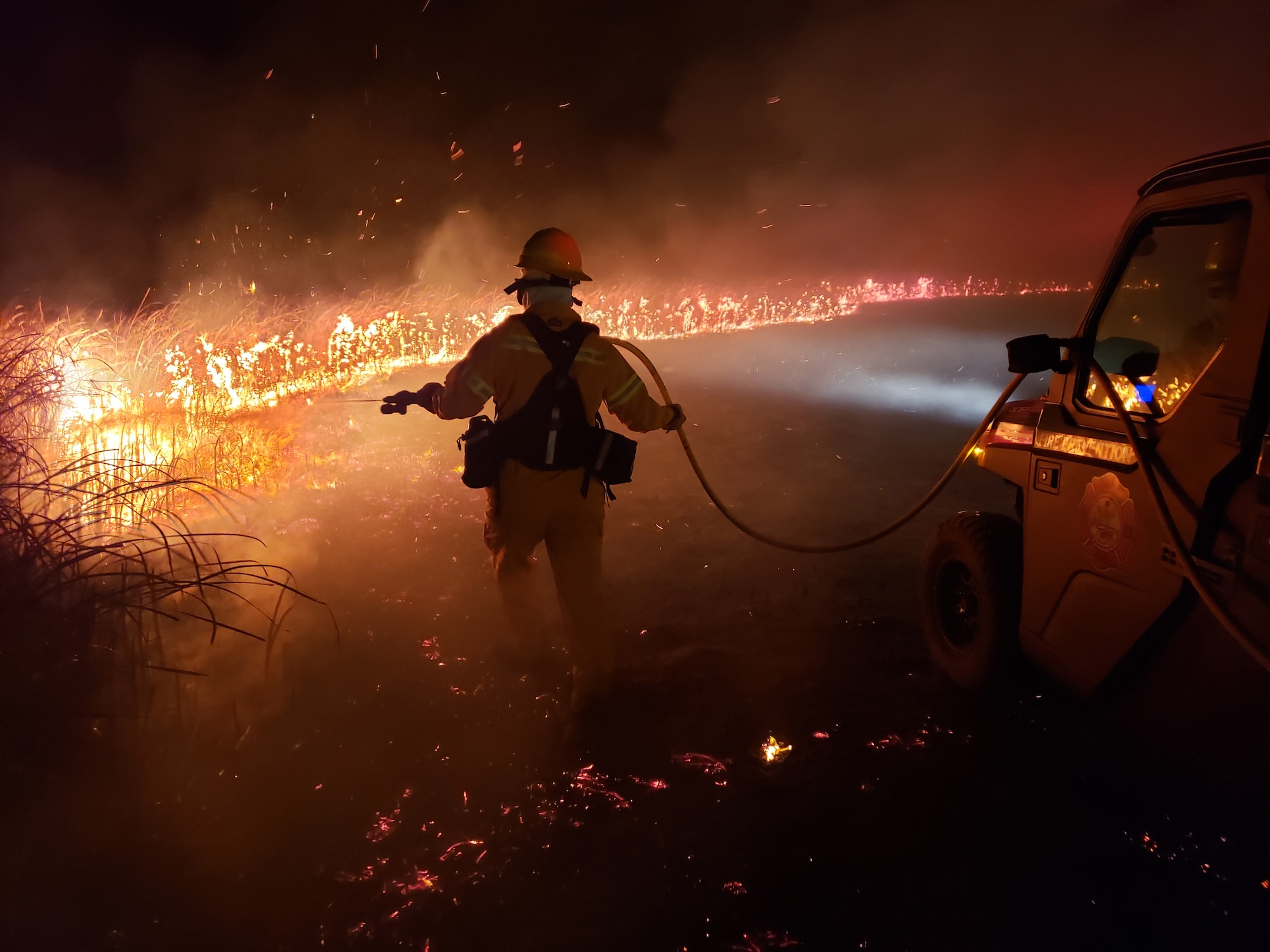 A firefighter wearing yellow sprays water on an orange wildland fire in the dark.