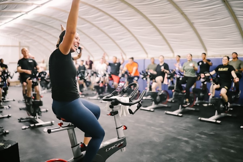 A woman wearing a headset sits on an exercise bike and gestures to a large group of people working out on bikes.