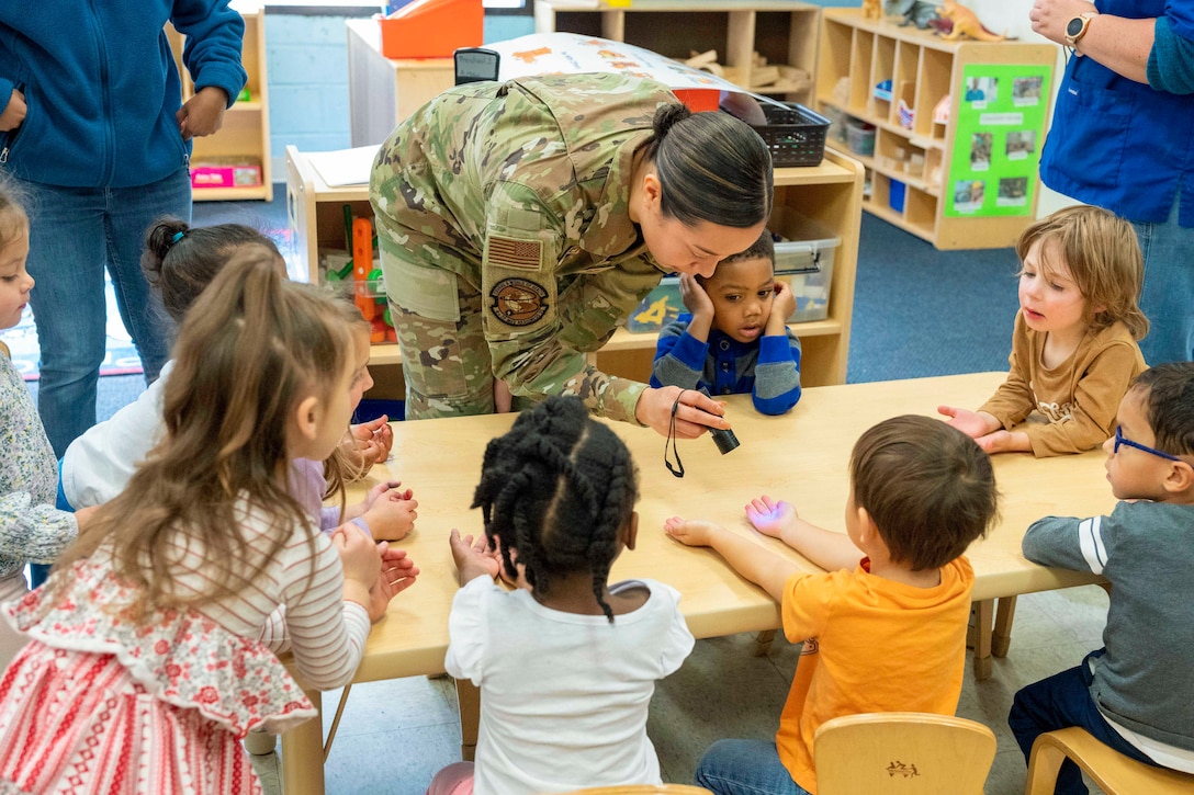 An airman uses a black light to check a child’s hands who is sitting a table with fellow students.