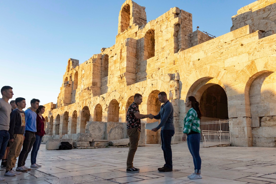 Two Marines shake hands in front of a group of students with an ancient Greek monument in the background.