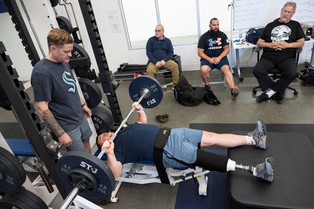An RSM with a right leg amputation bench presses while teammates and coaches look on.