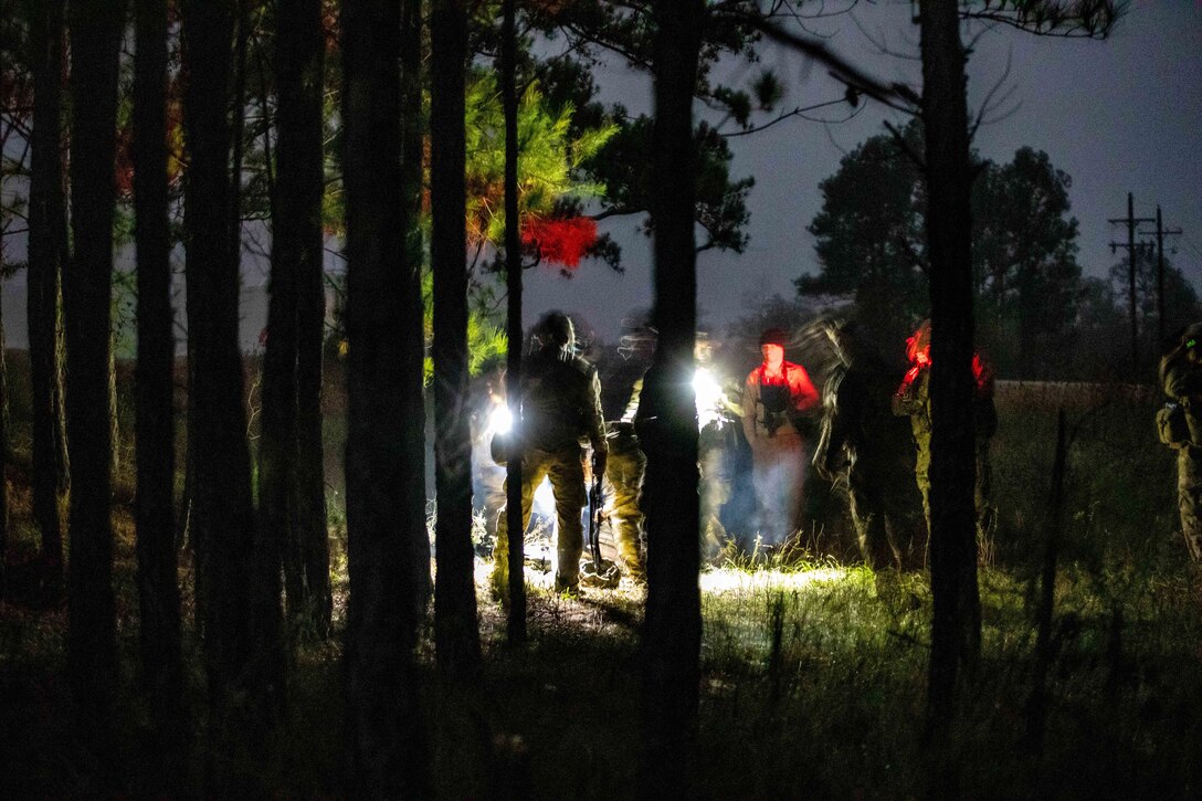 Soldiers gather in the woods in the dark illuminated by white and red lights.
