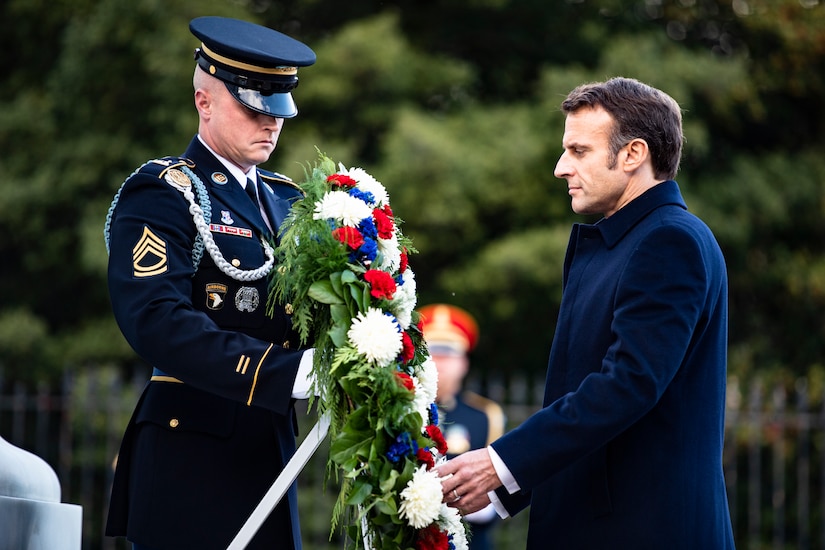 A man looks at a wreath being held by a service member in a dress uniform.