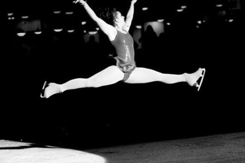 A woman figure skating leaps into the air with her legs horizontal and parallel to the ground.