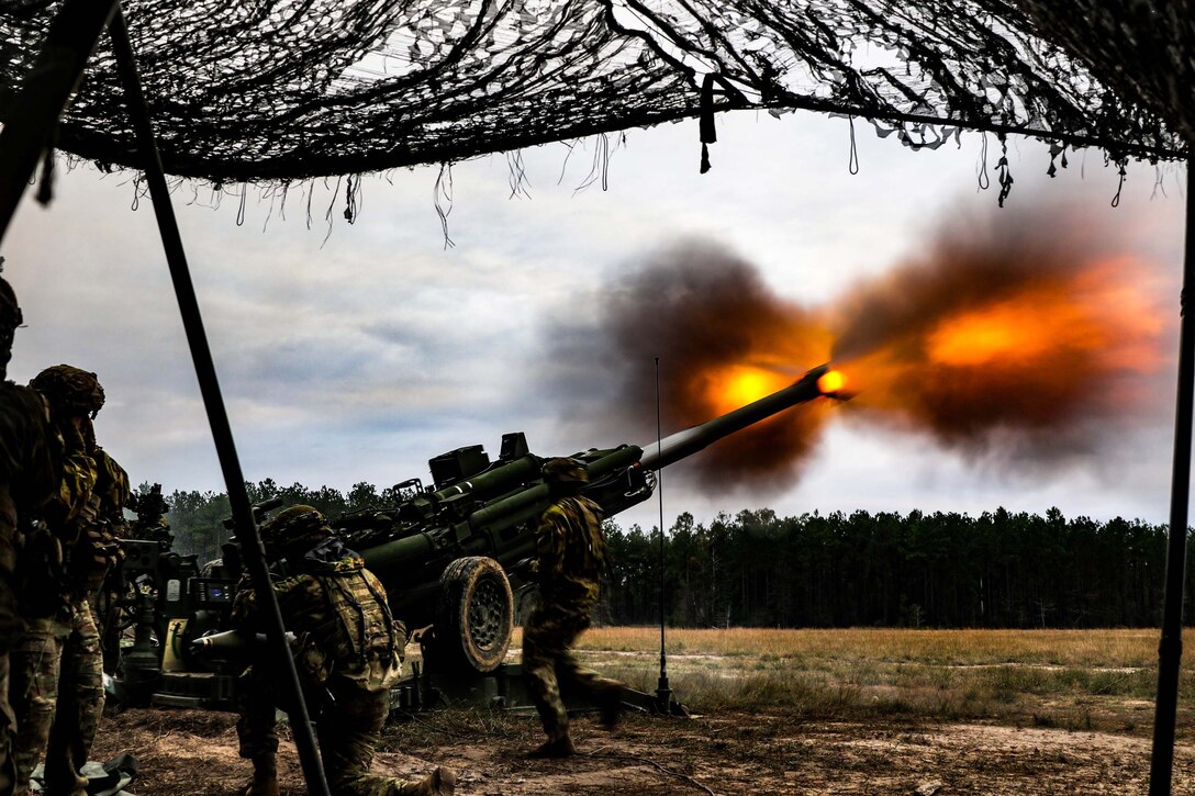 Two soldiers stand behind a large ground weapon shooting out fire.