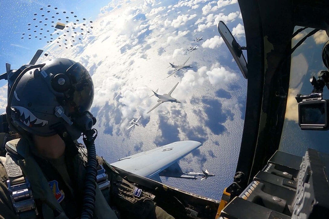 A pilot looks out a window at several planes below while flying above the clouds.