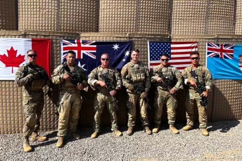 Six U.S. soldiers pose for a photo in front of multiple country flags displayed on a wall.