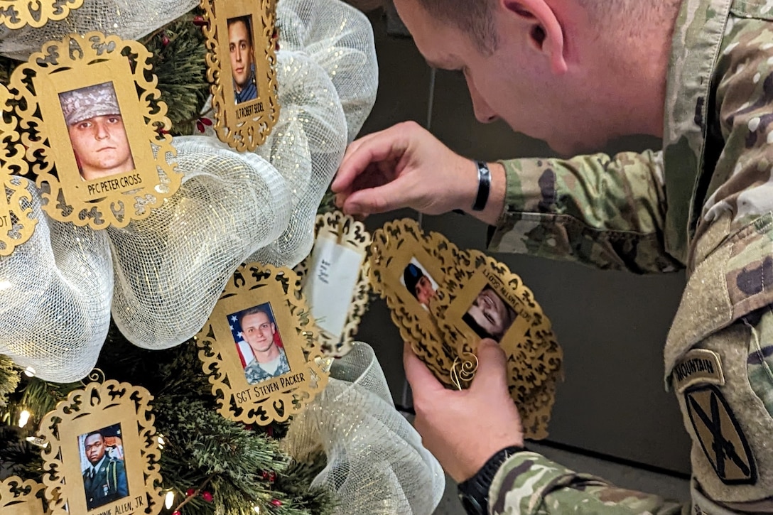 A soldier hangs ornaments of framed photos of soldiers on a tree.
