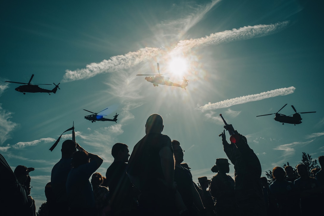 Silhouettes of people look upwards at helicopters flying overhead.