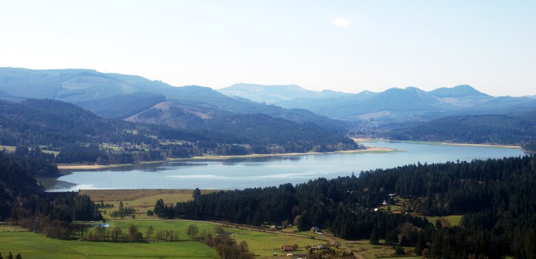 An aerial photograph shows a concrete dam from above, and a lake behind the dam, in a valley with green grass and hills of evergreen trees on a sunny, clear day with a perfectly blue sky.