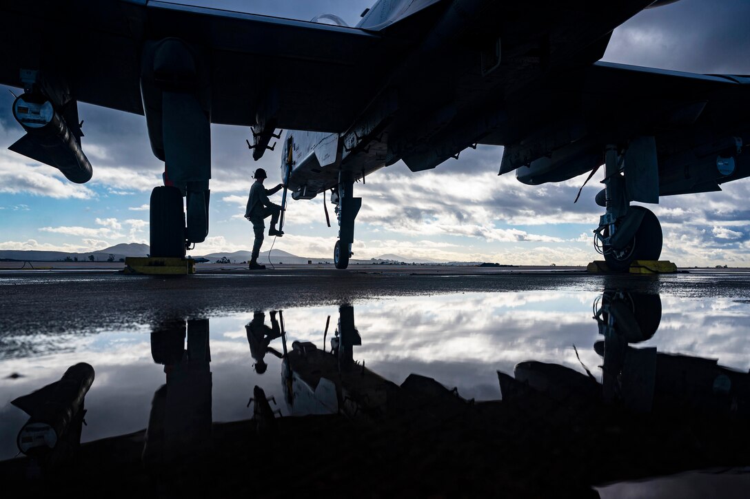 An airmen is reflected on the ground while climbing up to an aircraft in silhouette on a flight line.