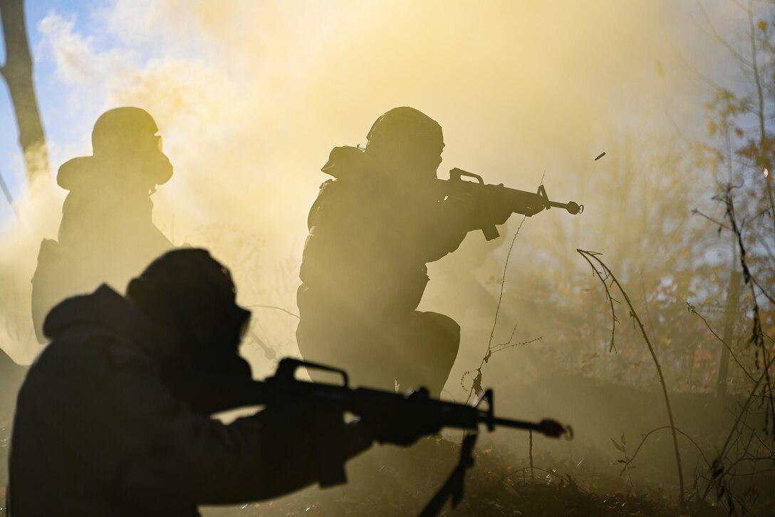 Airmen in protective gear shown in silhouette fire rifles outdoors amid a smoke cloud.