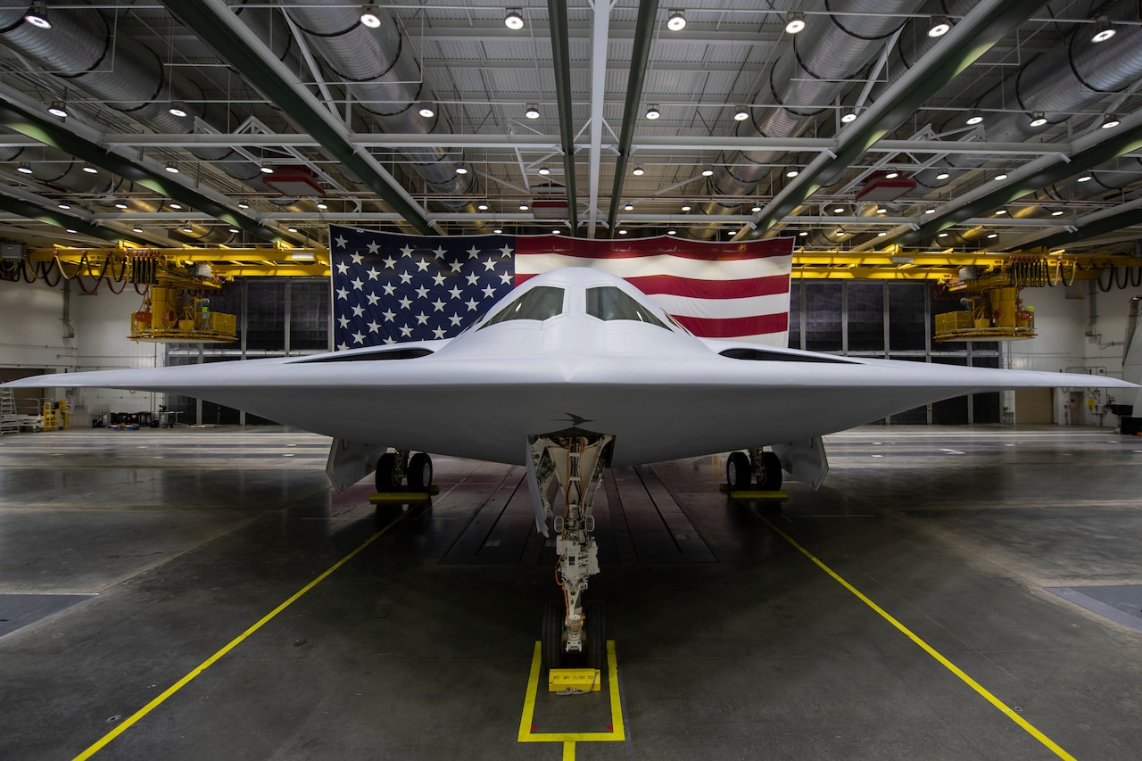 The B-21 raider aircraft sits in a hangar with the American flag in the background.