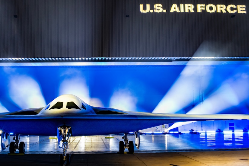 A B-21 Raider aircraft is unveiled with blue and white lights in the background.