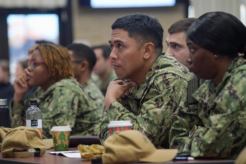 People in camouflage uniforms sit at a table looking straight ahead.