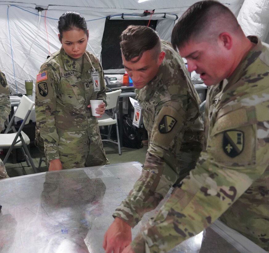 Three service members stand around a table; two of them point at a map.