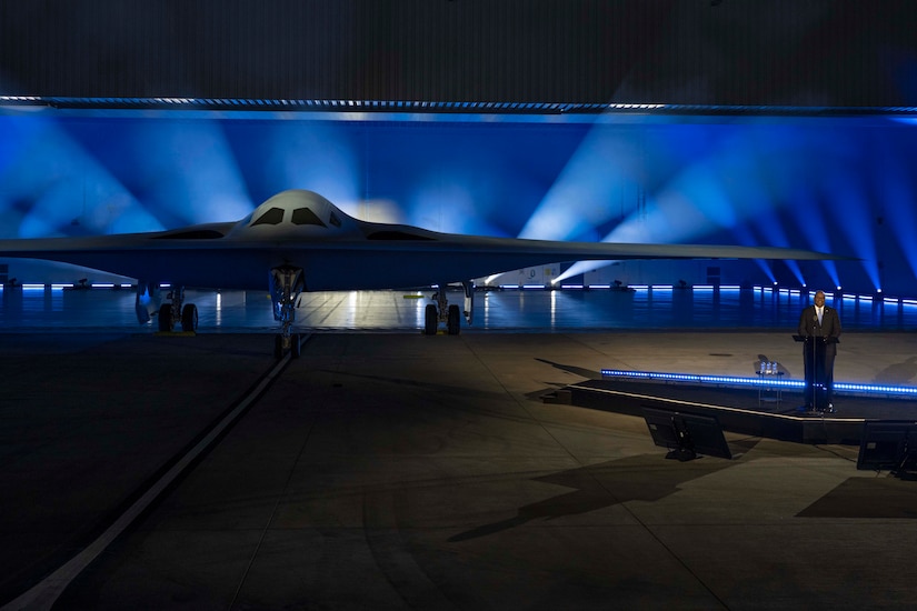 Secretary of Defense Lloyd J. Austin III stands behind a lectern next to an aircraft in a hangar.