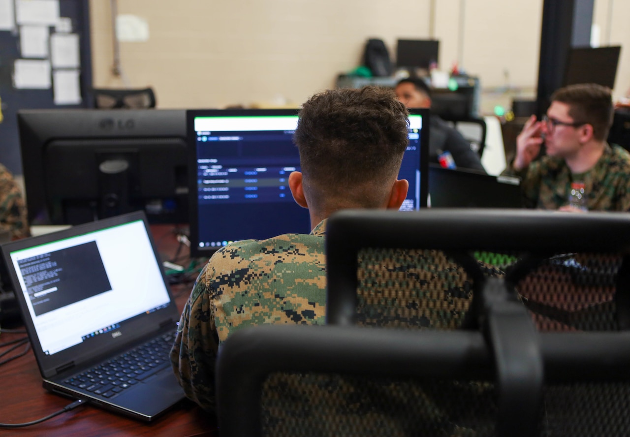 A group of Marines work at desks with computer monitors.