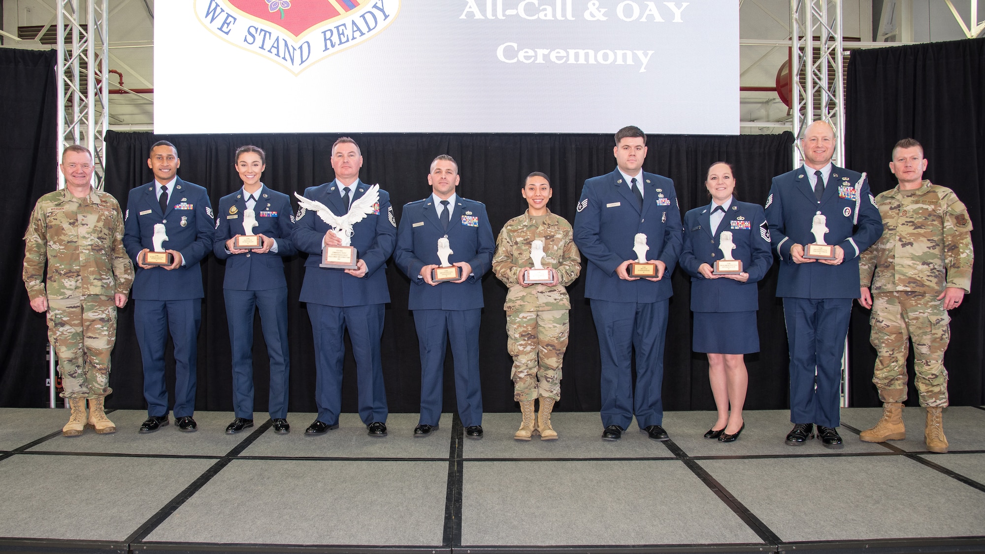 10 uniformed military members stand in a line, middle 8 are holding trophies.