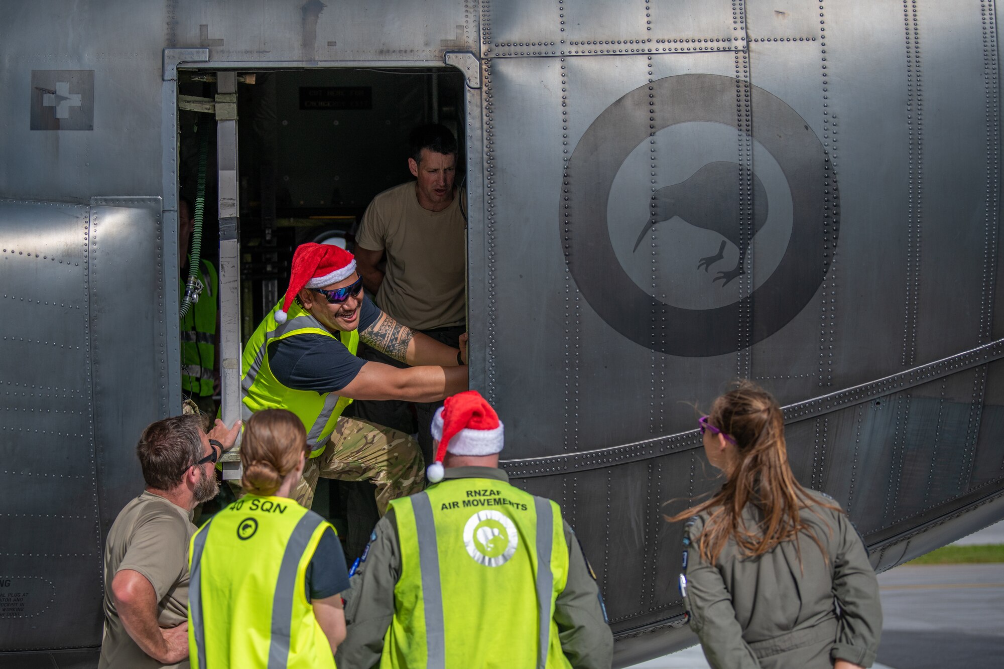 Members of the Royal New Zealand Air Force wear festive attire while hand cranking the loading ramp door of a C-130H Hercules close.
