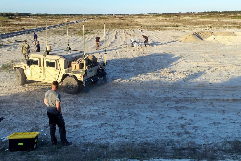 Service members work in a field with an unmanned aerial vehicle as a Humvee is parked nearby.