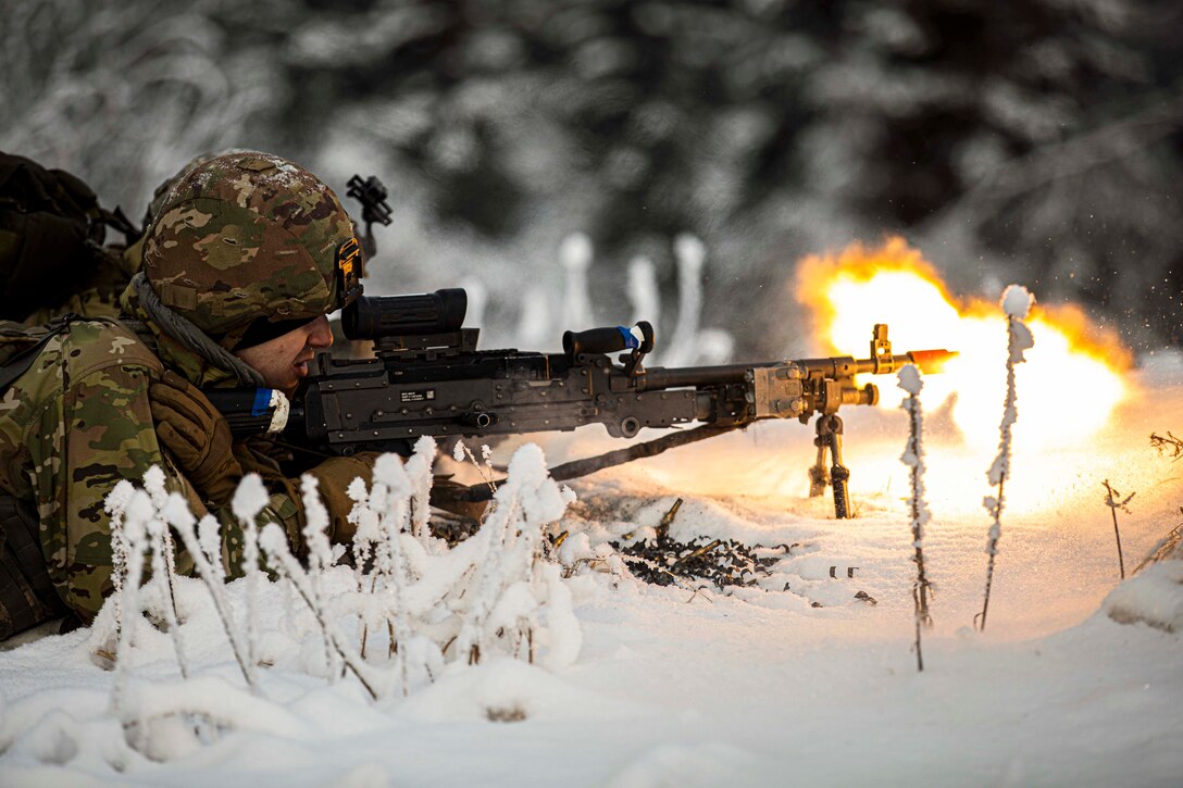 A National Guardsman fires a weapon while laying in the snow in the woods.