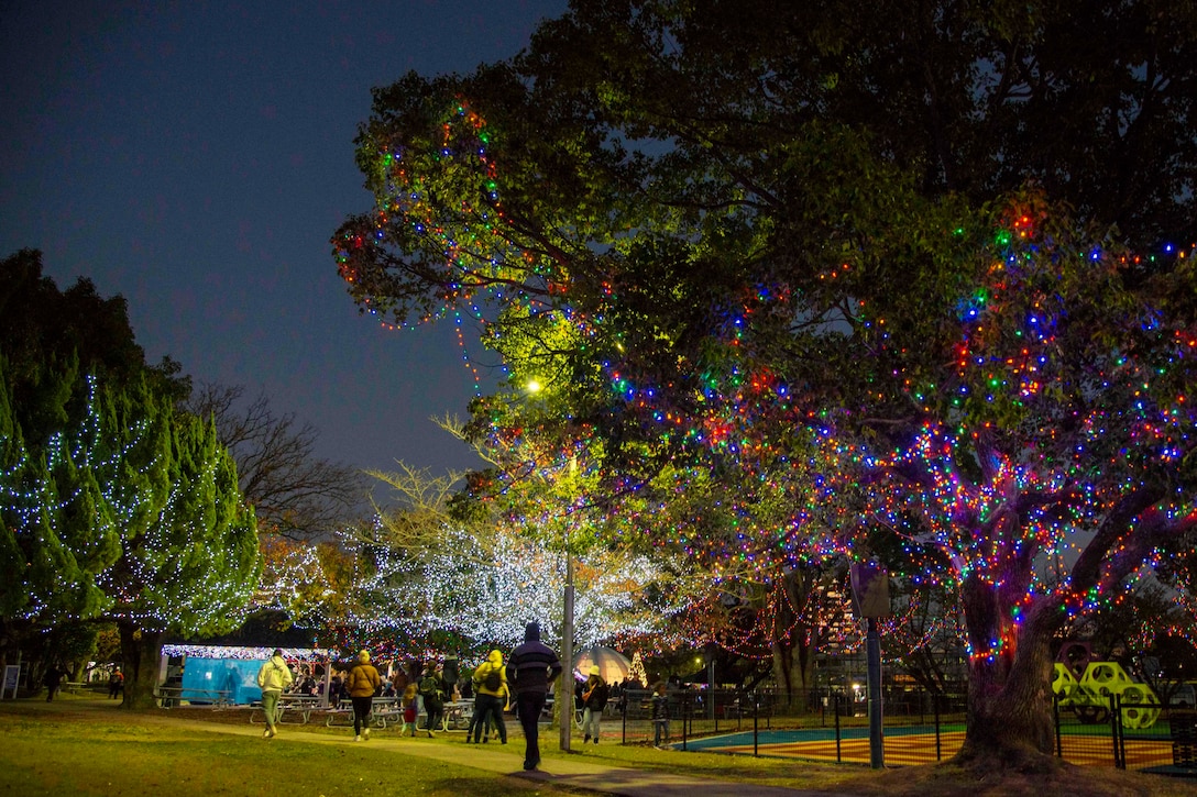 Sailor, family and community members walk in a park with tree covered in Christmas lights.
