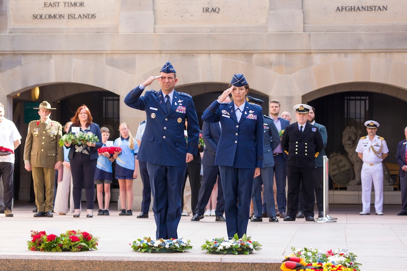 Two service members salute while standing in front of four wreaths on the ground ;  a group of people stands in the background.