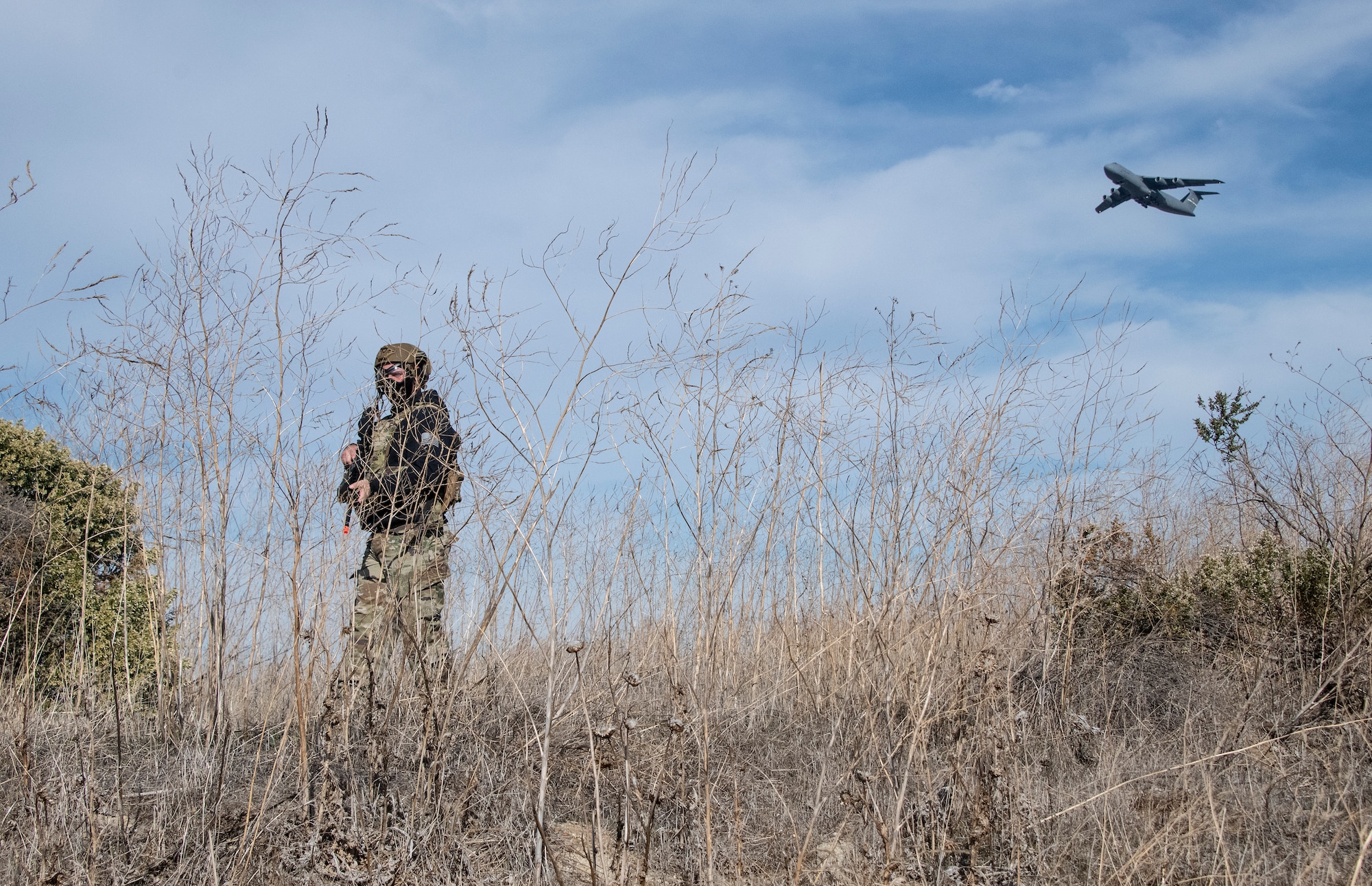 Airman walks through tall weeds while C-5 Super Galaxy files overhead.