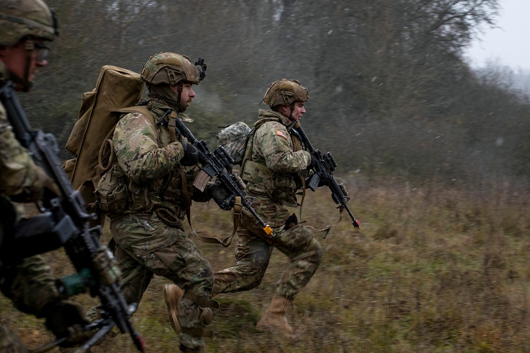 Three soldiers run over grass carrying rifles.