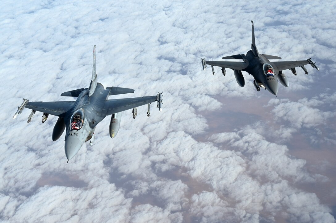 Two jets fly in cloud-dotted sky, one with the U.S. flag displayed in its cockpit and the other with the Dutch flag.