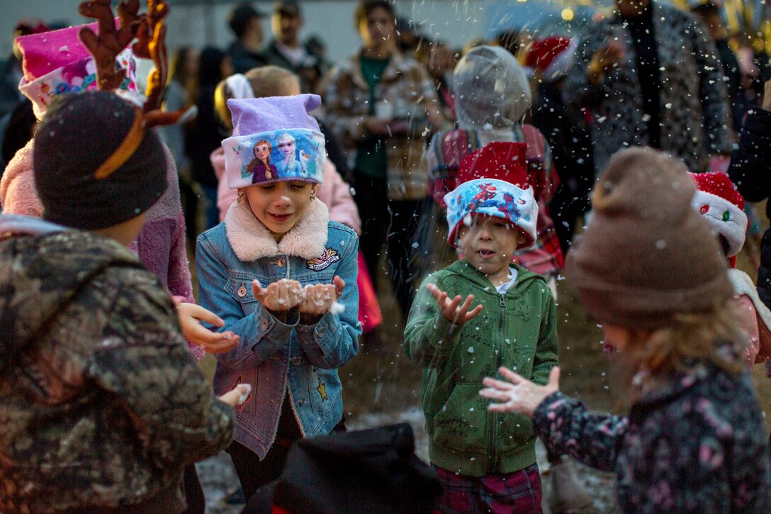 Children in holiday garb hold out their hands to touch fake snow falling outside.