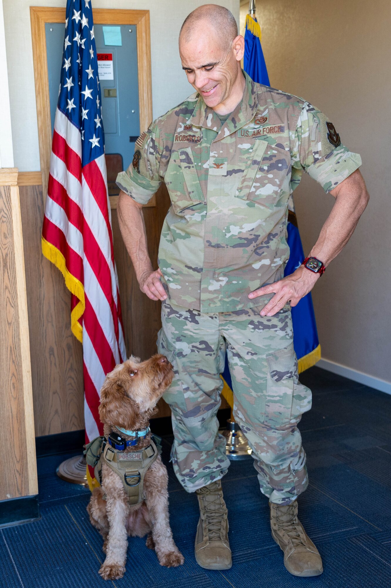 U.S. Air Force Col. Adam Roberts, the 555 RED HORSE Squadron commander, and his service dog, Porsche, pose in front of the American flag at Nellis Air Force base, Nevada, September 20, 2022. Roberts trained Porsche to function as a service animal and to be able to engage with fellow Airmen by accepting vulnerability. (U.S. Air Force photo by Staff Sgt. Timothy Leddick) (this photo has been edited to enhance the subjects)