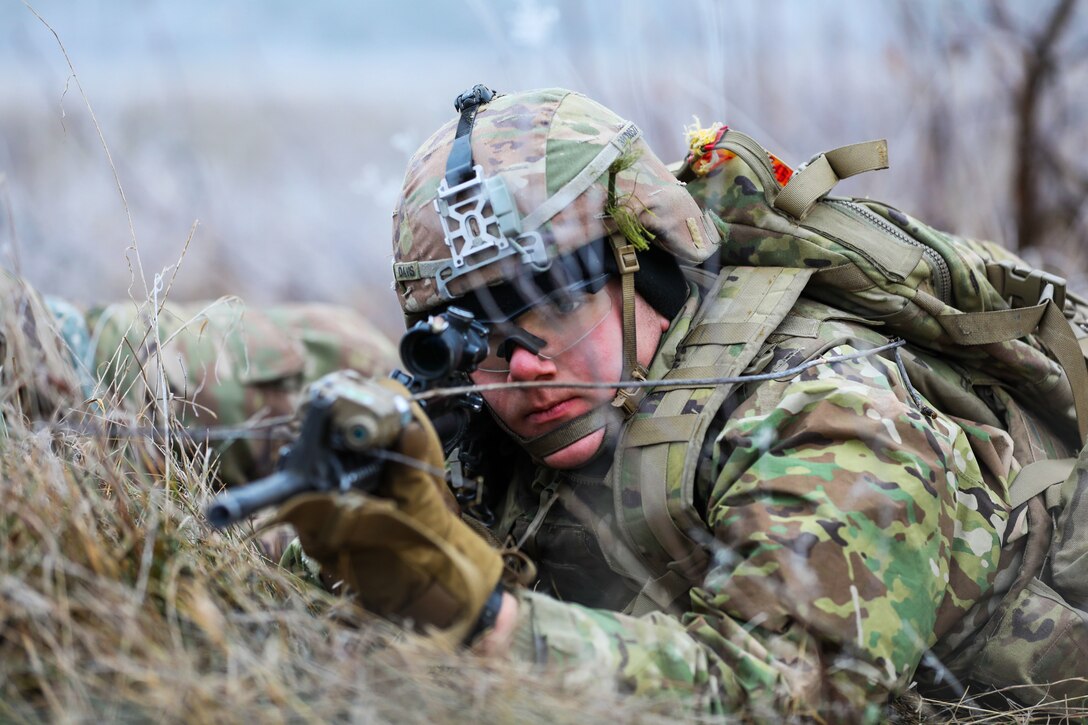 A soldier lays on the ground while looking through the scope of his weapon.