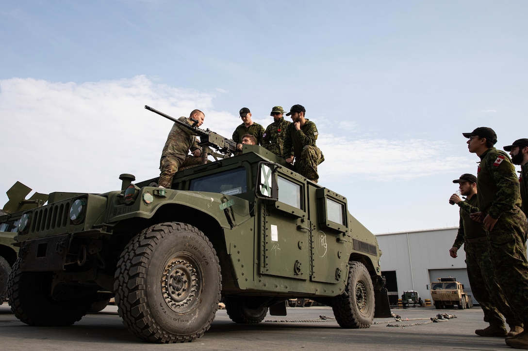 U.S. and Canadian soldiers on top of a vehicle look at the attached weapon while others look on from the ground.