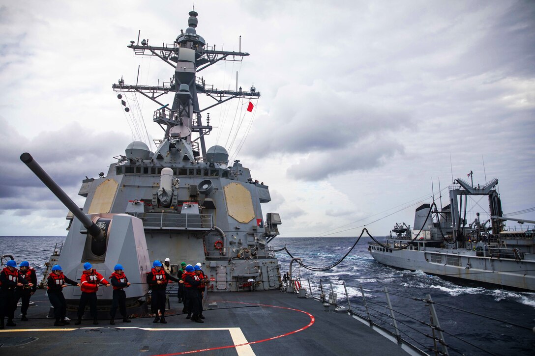 Sailors on a ship at sea hold a line during a replenishment with a nearby ship.